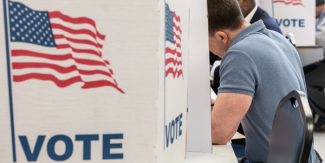 A voter sits at a booth marked with "VOTE" and an American flag, participating in an election at a polling station. A voter sits at a booth marked with "VOTE" and an American flag, participating in an election at a polling station.
